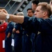 A military or naval personnel member in dark blue coveralls is aiming a semi-automatic pistol in a training or demonstration exercise on what appears to be the deck of a naval vessel. A group of similarly uniformed personnel stand in a line behind him, observing the activity. The setting and uniforms suggest a U.S. Navy or Coast Guard firearms qualification or safety training exercise.