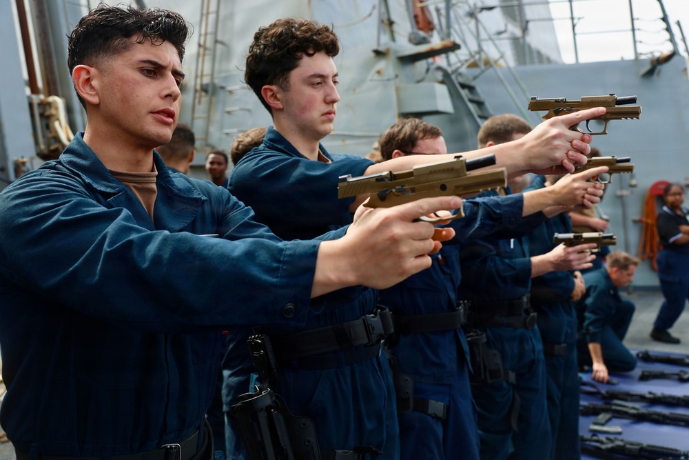 A group of military personnel in navy blue coveralls are conducting a firearms training exercise on the deck of a naval vessel. They are standing in a line, aiming tan/FDE-colored pistols in a coordinated drill. Additional weapons are visible laid out on a mat on the deck, suggesting a structured weapons familiarization or qualification exercise.