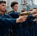 A group of military personnel in navy blue coveralls are conducting a firearms training exercise on the deck of a naval vessel. They are standing in a line, aiming tan/FDE-colored pistols in a coordinated drill. Additional weapons are visible laid out on a mat on the deck, suggesting a structured weapons familiarization or qualification exercise.
