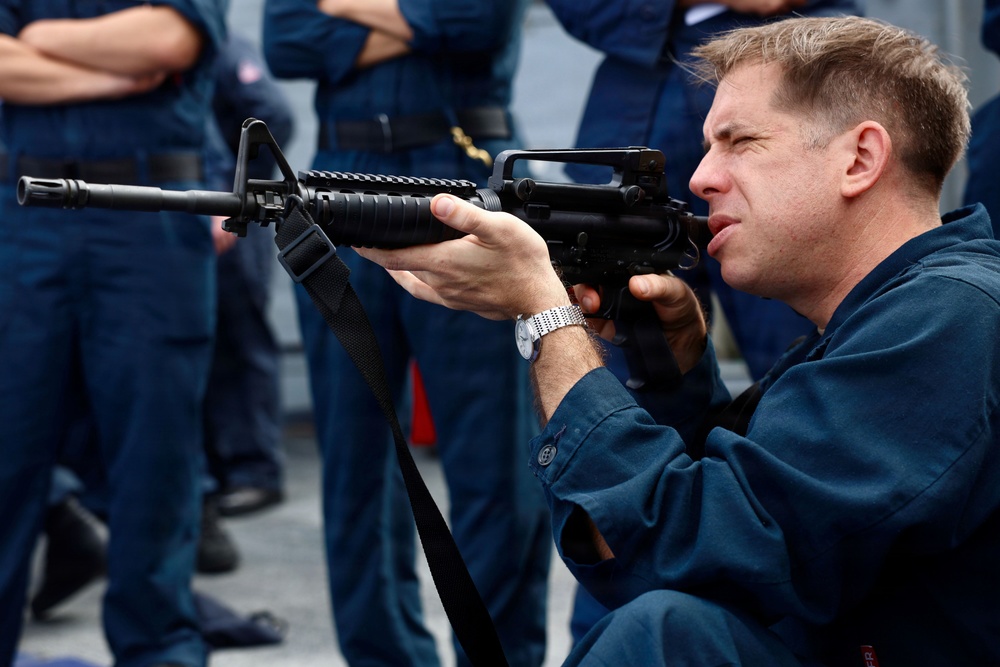 A man in dark blue military/naval coveralls is kneeling and aiming an M16/M4-style rifle in what appears to be a training or demonstration context. Several other personnel in similar dark blue uniforms are visible standing in the background. The setting appears to be on a ship's deck or similar military facility.