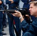 A man in dark blue military/naval coveralls is kneeling and aiming an M16/M4-style rifle in what appears to be a training or demonstration context. Several other personnel in similar dark blue uniforms are visible standing in the background. The setting appears to be on a ship's deck or similar military facility.