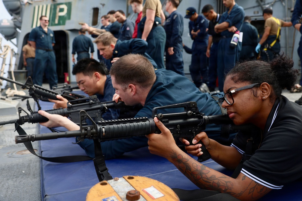 A group of individuals in what appears to be a military training exercise on the deck of a naval vessel. Three people in the foreground are in prone shooting positions holding M16/M4-style rifles, while numerous uniformed personnel observe in the background. The setting appears to be a formal weapons familiarization or qualification drill.
