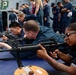 A group of individuals in what appears to be a military training exercise on the deck of a naval vessel. Three people in the foreground are in prone shooting positions holding M16/M4-style rifles, while numerous uniformed personnel observe in the background. The setting appears to be a formal weapons familiarization or qualification drill.