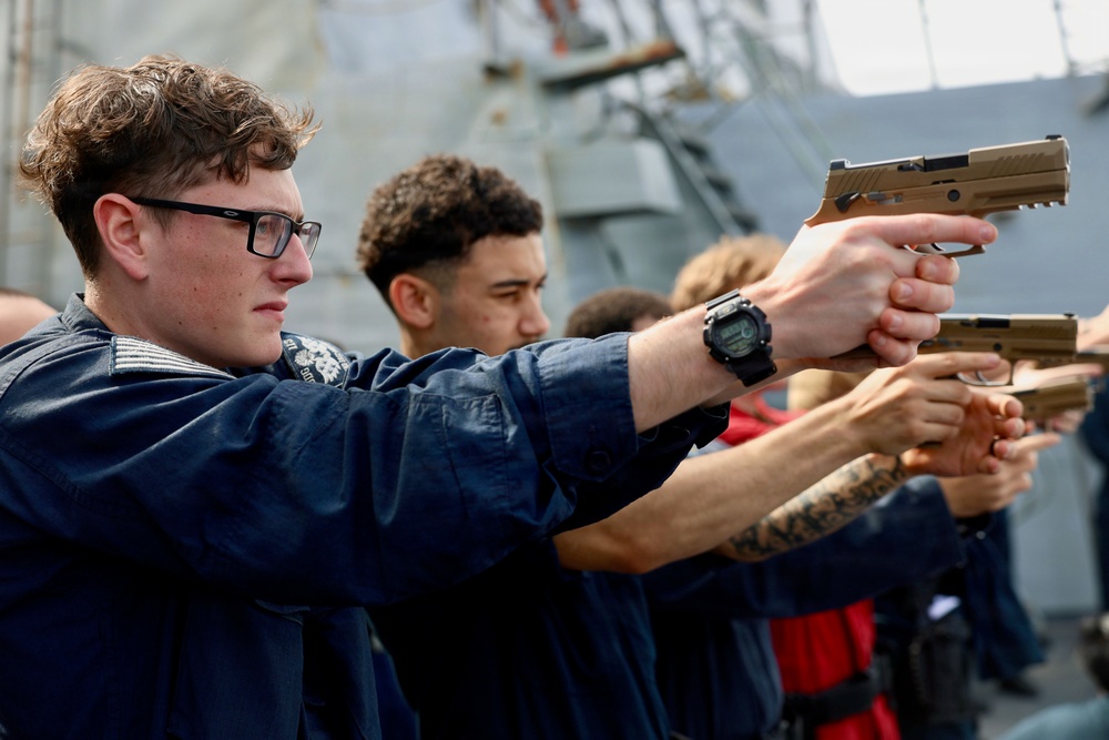 A group of individuals in dark blue uniforms are participating in what appears to be a firearms training exercise, aiming tan/coyote-colored pistols in a line formation. The setting appears to be on the deck of a naval vessel, visible from the gray metal superstructure in the background. The exercise resembles a military or coast guard qualification drill.