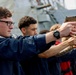A group of individuals in dark blue uniforms are participating in what appears to be a firearms training exercise, aiming tan/coyote-colored pistols in a line formation. The setting appears to be on the deck of a naval vessel, visible from the gray metal superstructure in the background. The exercise resembles a military or coast guard qualification drill.
