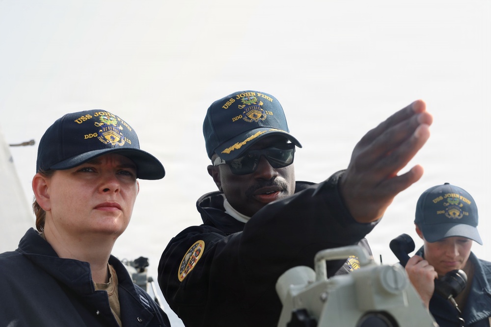 Three U.S. Navy personnel are photographed on the deck of USS John Finn (DDG-113), a guided-missile destroyer. A senior sailor appears to be pointing and giving instruction or direction to a junior sailor, while a third crew member holds a radio or communication device in the background. All three are wearing USS John Finn ship's company ball caps.