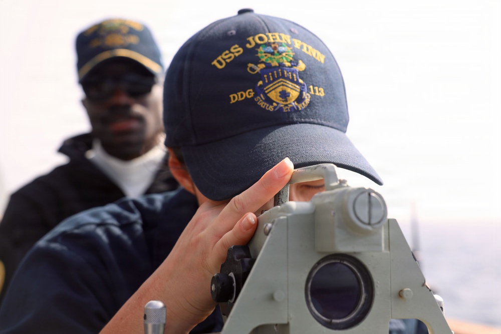 Two naval personnel are shown on the deck of a U.S. Navy ship. The foreground figure is using a navigational sighting instrument, wearing a cap embroidered with 'USS John Finn DDG 113'. A second sailor in the background observes the activity.