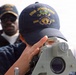 Two naval personnel are shown on the deck of a U.S. Navy ship. The foreground figure is using a navigational sighting instrument, wearing a cap embroidered with 'USS John Finn DDG 113'. A second sailor in the background observes the activity.