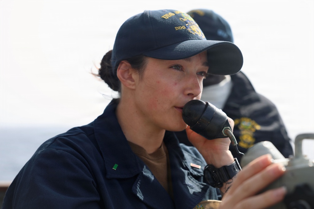 A person in U.S. Navy uniform is speaking into a handheld radio/microphone while aboard what appears to be a naval vessel. They are wearing a navy blue cap with 'DDG' visible and gold embroidery, consistent with a U.S. Navy destroyer crew cap. A second uniformed individual is visible in the background.