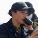 A person in U.S. Navy uniform is speaking into a handheld radio/microphone while aboard what appears to be a naval vessel. They are wearing a navy blue cap with 'DDG' visible and gold embroidery, consistent with a U.S. Navy destroyer crew cap. A second uniformed individual is visible in the background.