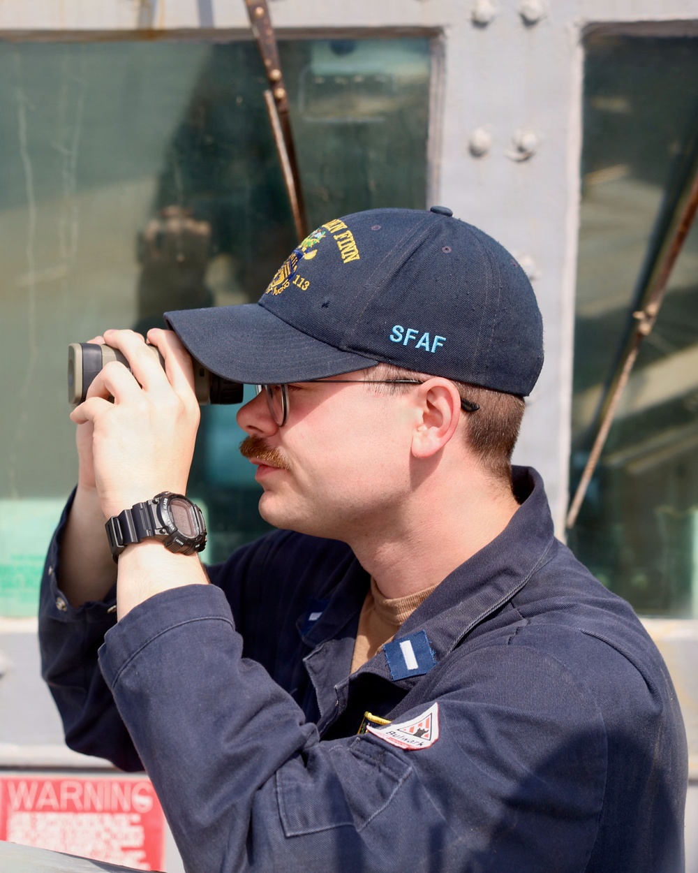 A male individual wearing a navy blue uniform and cap is looking through binoculars or a rangefinder device. He is positioned in front of what appears to be a military or naval vessel structure with metal plating and windows visible in the background. His cap displays 'SFAF' in teal lettering and an emblem with the number 113.