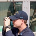 A male individual wearing a navy blue uniform and cap is looking through binoculars or a rangefinder device. He is positioned in front of what appears to be a military or naval vessel structure with metal plating and windows visible in the background. His cap displays 'SFAF' in teal lettering and an emblem with the number 113.