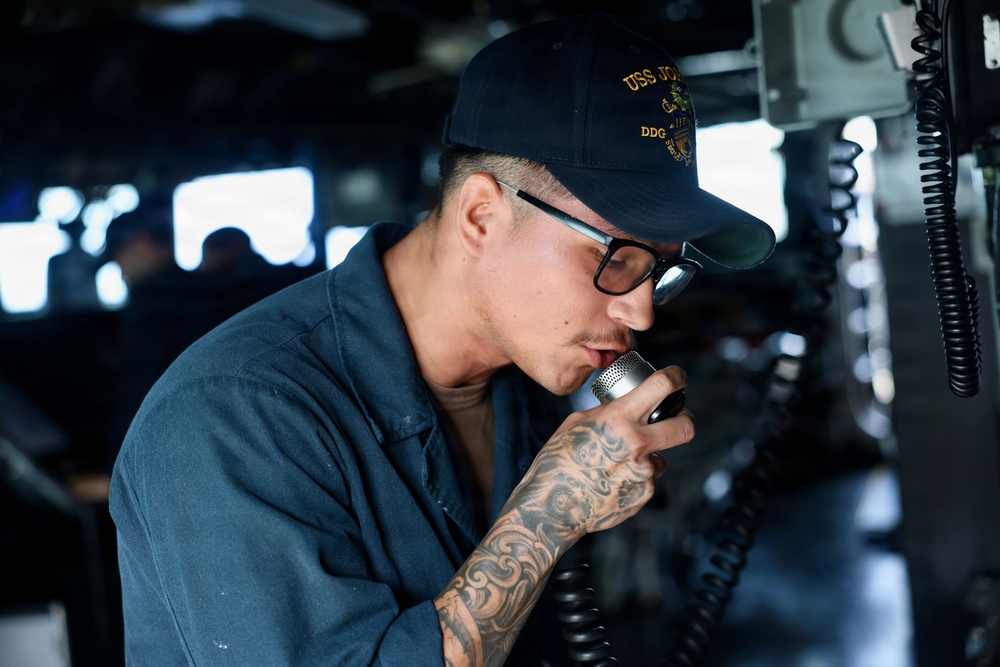 A U.S. Navy sailor is speaking into a shipboard communication handset, likely making an announcement or relaying information. The sailor is wearing a navy blue uniform and a USS ship cap with 'DDG' designation visible, indicating assignment to a guided-missile destroyer. The setting appears to be the bridge or a communications station aboard a naval vessel.