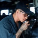 A U.S. Navy sailor is speaking into a shipboard communication handset, likely making an announcement or relaying information. The sailor is wearing a navy blue uniform and a USS ship cap with 'DDG' designation visible, indicating assignment to a guided-missile destroyer. The setting appears to be the bridge or a communications station aboard a naval vessel.