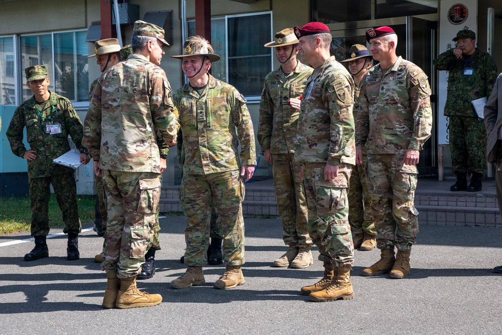 A group of multinational military personnel are gathered outdoors in front of a building, appearing to be engaged in informal conversation. The group includes soldiers wearing different uniform styles and headgear, suggesting a joint or combined military exercise or meeting. Japanese Ground Self-Defense Force personnel are visible alongside what appear to be U.S. Army and Australian Defence Force soldiers.