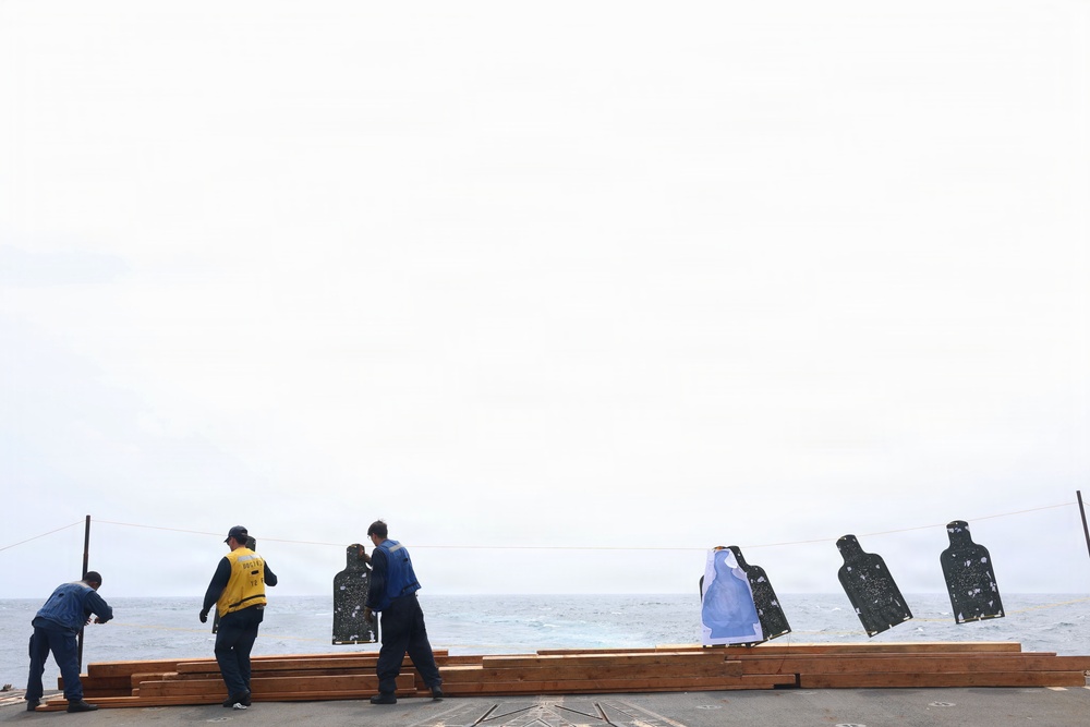 The image shows several individuals on what appears to be the deck of a naval vessel at sea, setting up or handling shooting range targets. Multiple human silhouette targets are suspended on a line, some showing visible bullet hole patterns. The overcast sky and open ocean are visible in the background.