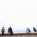 The image shows several individuals on what appears to be the deck of a naval vessel at sea, setting up or handling shooting range targets. Multiple human silhouette targets are suspended on a line, some showing visible bullet hole patterns. The overcast sky and open ocean are visible in the background.