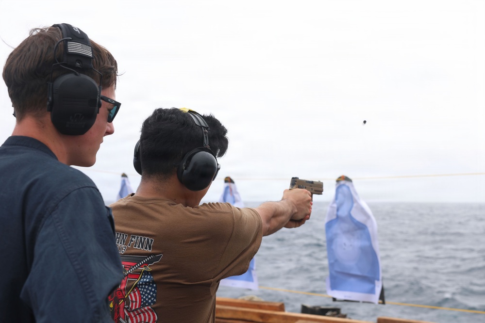 Two individuals wearing hearing protection are engaged in a pistol shooting exercise on what appears to be the deck of a naval vessel at sea. One person is actively firing a tan/FDE-colored semi-automatic pistol toward paper silhouette targets set up along a rope line over open water. A spent cartridge casing is visibly in the air near the muzzle of the firearm.