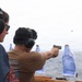 Two individuals wearing hearing protection are engaged in a pistol shooting exercise on what appears to be the deck of a naval vessel at sea. One person is actively firing a tan/FDE-colored semi-automatic pistol toward paper silhouette targets set up along a rope line over open water. A spent cartridge casing is visibly in the air near the muzzle of the firearm.