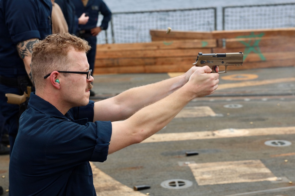 A male service member in dark navy blue uniform is firing a pistol on what appears to be the deck of a naval vessel. He is wearing ear protection (foam earplugs) and glasses, using a two-handed grip on a tan/FDE-colored semi-automatic pistol. A spent casing is visible ejecting from the firearm, and other personnel are visible observing in the background.