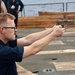 A male service member in dark navy blue uniform is firing a pistol on what appears to be the deck of a naval vessel. He is wearing ear protection (foam earplugs) and glasses, using a two-handed grip on a tan/FDE-colored semi-automatic pistol. A spent casing is visible ejecting from the firearm, and other personnel are visible observing in the background.