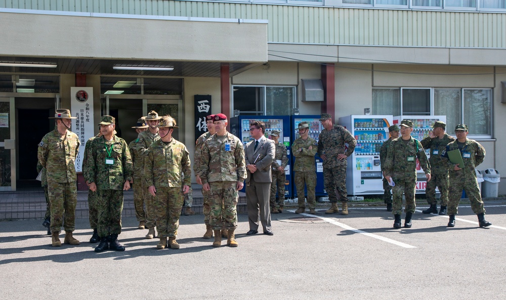 A group of military personnel from multiple nations are assembled outside a building with Japanese signage, suggesting a joint military exercise or briefing. Personnel are wearing various camouflage uniforms including what appear to be US Army, US Marine Corps, and Japan Ground Self-Defense Force uniforms, along with one individual in a civilian suit. The setting appears to be a Japanese military installation, consistent with a bilateral or multinational training exercise.
