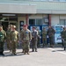 A group of military personnel from multiple nations are assembled outside a building with Japanese signage, suggesting a joint military exercise or briefing. Personnel are wearing various camouflage uniforms including what appear to be US Army, US Marine Corps, and Japan Ground Self-Defense Force uniforms, along with one individual in a civilian suit. The setting appears to be a Japanese military installation, consistent with a bilateral or multinational training exercise.