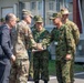 A multinational military gathering is taking place outdoors near a building, with personnel from at least three different armed forces present. A formal handshake is occurring between military officers, suggesting an official meeting or ceremony. A civilian in a business suit is also present among the uniformed personnel.