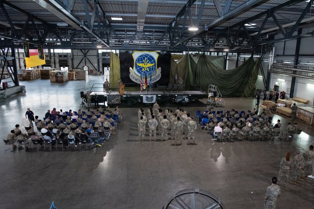 A military ceremony is taking place inside a large aircraft hangar, with personnel in camouflage uniforms arranged in formation and seated in chairs facing a raised stage. The backdrop features the Air Mobility Command insignia — a globe with wings — displayed prominently behind a podium. The event appears to be a formal military function such as a change of command, award ceremony, or unit activation.