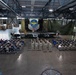 A military ceremony is taking place inside a large aircraft hangar, with personnel in camouflage uniforms arranged in formation and seated in chairs facing a raised stage. The backdrop features the Air Mobility Command insignia — a globe with wings — displayed prominently behind a podium. The event appears to be a formal military function such as a change of command, award ceremony, or unit activation.