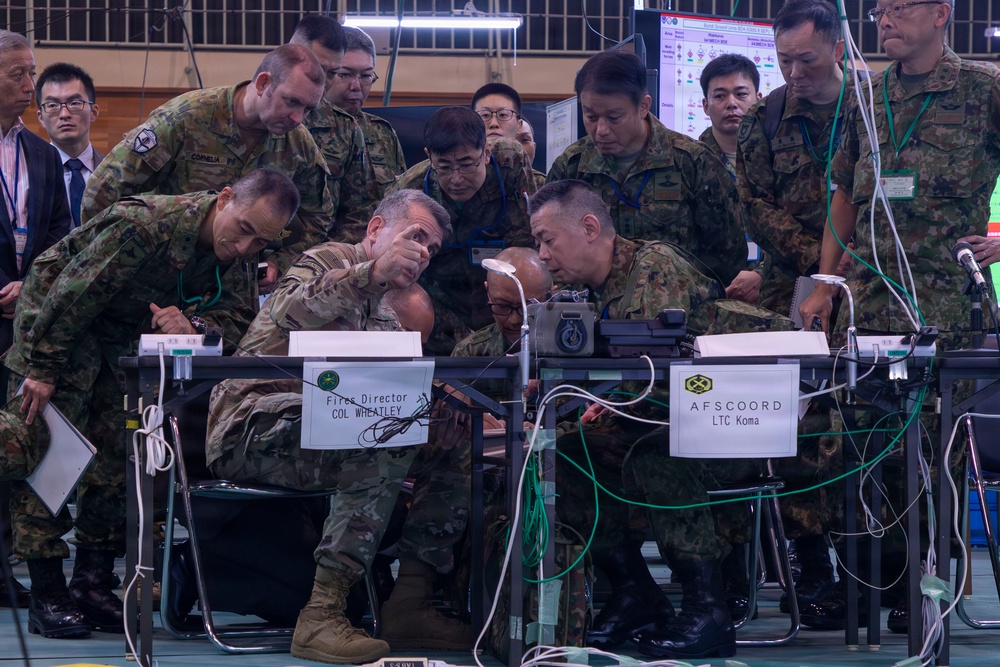 A joint military exercise or command post operation involving U.S. Army and Japanese Ground Self-Defense Force (JGSDF) personnel gathered around workstations. Name placards identify 'Fires Director COL Wheatley' and 'AFSCOORD LTC Koma,' indicating a combined fires coordination exercise. Personnel are closely examining equipment and documents at a command operations center setup.