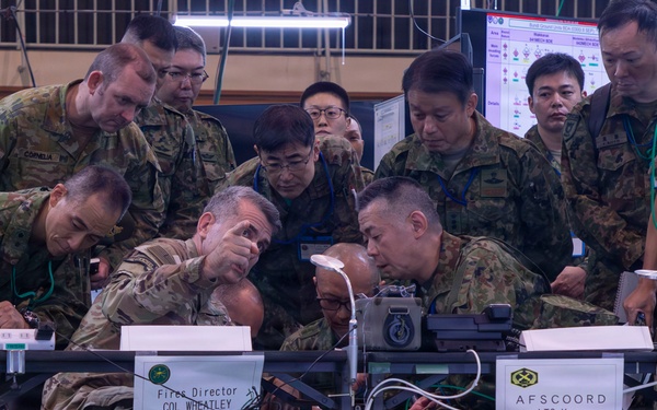 A joint military exercise or command post operation involving U.S. Army and Japanese Ground Self-Defense Force (JGSDF) personnel gathered around workstations. Name placards identify 'Fires Director COL Wheatley' and 'AFSCOORD LTC Koma,' indicating a combined fires coordination exercise. Personnel are closely examining equipment and documents at a command operations center setup.