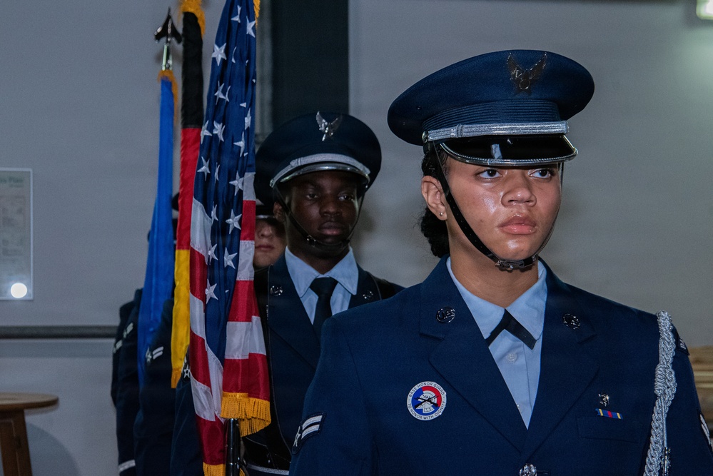 The image shows members of a military color guard in formal dress uniforms, likely U.S. Air Force, standing at attention during a ceremony. They are carrying flags including the American flag and at least one other flag with gold fringe. The foreground figure wears a distinctive honor guard patch and silver aiguillette on their uniform.