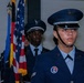 The image shows members of a military color guard in formal dress uniforms, likely U.S. Air Force, standing at attention during a ceremony. They are carrying flags including the American flag and at least one other flag with gold fringe. The foreground figure wears a distinctive honor guard patch and silver aiguillette on their uniform.