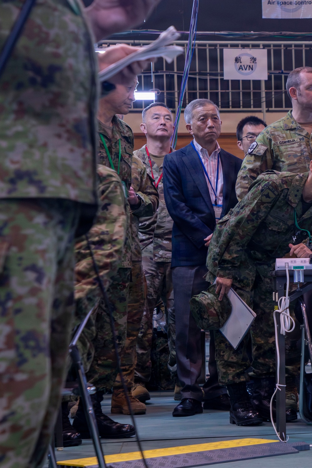 A group of military personnel and civilian officials are gathered in what appears to be a command or operations center, likely during a joint military exercise or briefing. Multiple nationalities are represented, including Japanese Ground Self-Defense Force soldiers and at least one Western military officer with a name tape reading 'CORNELIA'. Civilians wearing lanyards are present alongside uniformed personnel, suggesting an official observation or coordination event.