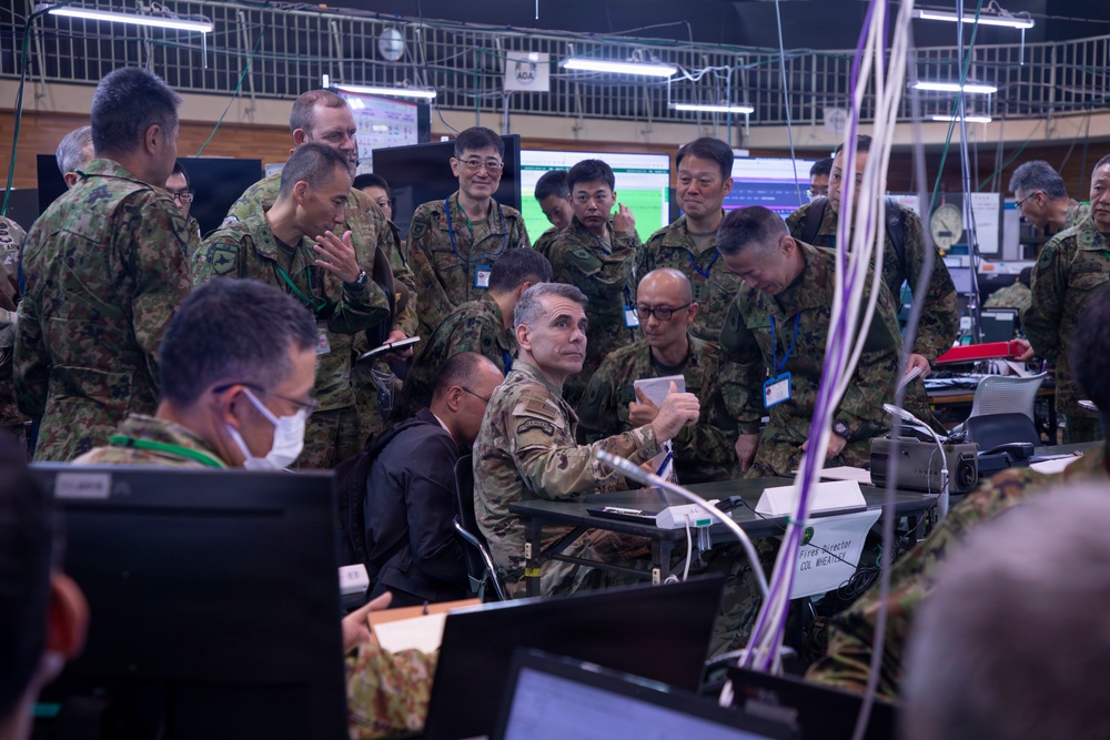 A joint military operations center showing personnel from what appears to be the U.S. Army and Japan Ground Self-Defense Force (JGSDF) gathered around workstations during what appears to be a combined exercise or briefing. A U.S. Army officer in MultiCam uniform is seated at a desk marked 'Fires Director COL WHEATLEY' while surrounded by numerous JGSDF personnel in Japanese camouflage uniforms. The setting is a command and control facility with multiple computer monitors, cables, and communications equipment.
