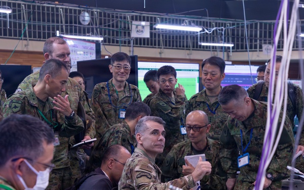 A joint military operations center showing personnel from what appears to be the U.S. Army and Japan Ground Self-Defense Force (JGSDF) gathered around workstations during what appears to be a combined exercise or briefing. A U.S. Army officer in MultiCam uniform is seated at a desk marked 'Fires Director COL WHEATLEY' while surrounded by numerous JGSDF personnel in Japanese camouflage uniforms. The setting is a command and control facility with multiple computer monitors, cables, and communications equipment.