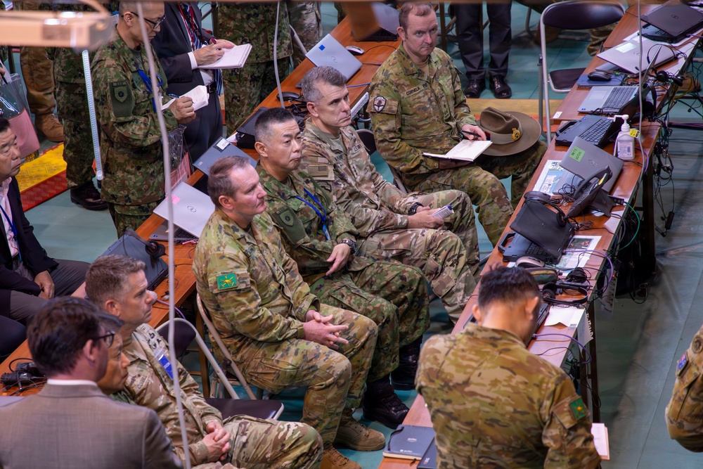 A multinational military briefing or command post exercise is underway in what appears to be an operations center or conference hall. Personnel from multiple nations — identifiable by distinct camouflage patterns and insignia including Australian Army (AUSCAM), Japanese Ground Self-Defense Force, and U.S. Army uniforms — are seated and standing around tables equipped with laptops and communications equipment. The group appears to be attentively observing a presentation or briefing occurring off-camera.