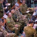 A multinational military briefing or command post exercise is underway in what appears to be an operations center or conference hall. Personnel from multiple nations — identifiable by distinct camouflage patterns and insignia including Australian Army (AUSCAM), Japanese Ground Self-Defense Force, and U.S. Army uniforms — are seated and standing around tables equipped with laptops and communications equipment. The group appears to be attentively observing a presentation or briefing occurring off-camera.