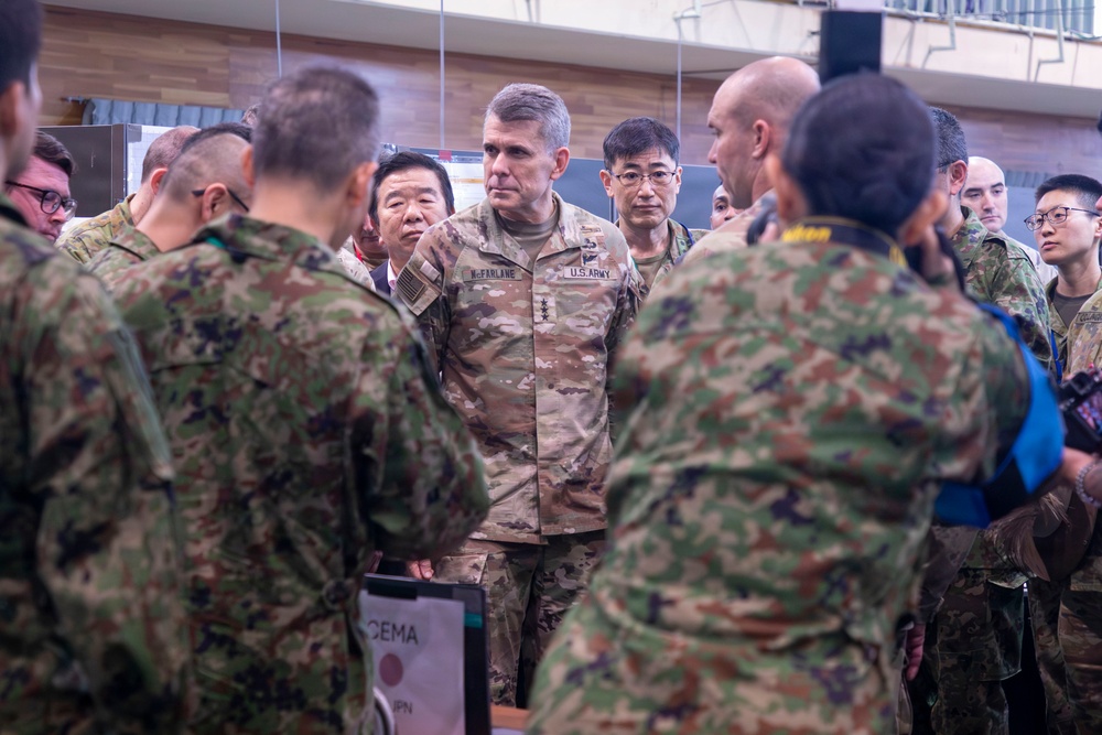 A U.S. Army officer, identifiable by the name tape 'McFARLANE' and 'U.S. ARMY' patch wearing what appears to be lieutenant general rank insignia, is engaged in a briefing or discussion with a mixed group of military personnel. The group includes soldiers in U.S. Army OCP (Operational Camouflage Pattern) uniforms and what appear to be Japanese Ground Self-Defense Force (JGSDF) soldiers in their distinctive camouflage pattern. The setting appears to be an indoor military facility, suggesting a joint military exercise or coordination meeting.