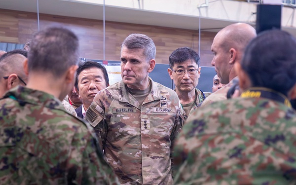 A U.S. Army officer, identifiable by the name tape 'McFARLANE' and 'U.S. ARMY' patch wearing what appears to be lieutenant general rank insignia, is engaged in a briefing or discussion with a mixed group of military personnel. The group includes soldiers in U.S. Army OCP (Operational Camouflage Pattern) uniforms and what appear to be Japanese Ground Self-Defense Force (JGSDF) soldiers in their distinctive camouflage pattern. The setting appears to be an indoor military facility, suggesting a joint military exercise or coordination meeting.