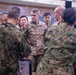 A U.S. Army officer, identifiable by the name tape 'McFARLANE' and 'U.S. ARMY' patch wearing what appears to be lieutenant general rank insignia, is engaged in a briefing or discussion with a mixed group of military personnel. The group includes soldiers in U.S. Army OCP (Operational Camouflage Pattern) uniforms and what appear to be Japanese Ground Self-Defense Force (JGSDF) soldiers in their distinctive camouflage pattern. The setting appears to be an indoor military facility, suggesting a joint military exercise or coordination meeting.