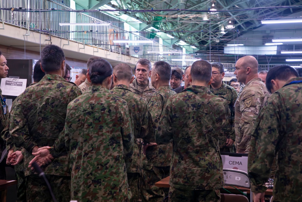 A group of military personnel from at least two different nations are gathered in what appears to be an indoor operations or briefing facility. The group includes soldiers wearing Japanese Ground Self-Defense Force (JGSDF) woodland camouflage uniforms alongside what appear to be U.S. military personnel in MultiCam/OCP uniforms. The gathering appears to be a joint military coordination meeting or briefing session.