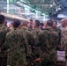 A group of military personnel from at least two different nations are gathered in what appears to be an indoor operations or briefing facility. The group includes soldiers wearing Japanese Ground Self-Defense Force (JGSDF) woodland camouflage uniforms alongside what appear to be U.S. military personnel in MultiCam/OCP uniforms. The gathering appears to be a joint military coordination meeting or briefing session.