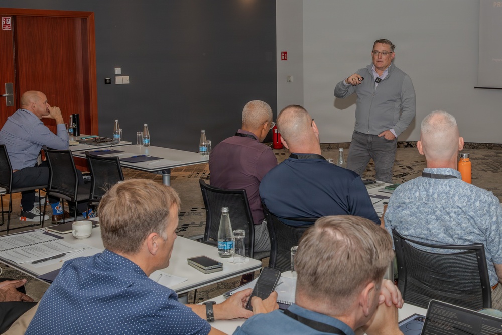 A professional training or business meeting is taking place in a modern conference room. A standing presenter in a gray sweater is addressing a group of approximately 7-8 seated male attendees arranged around white tables. The setting appears to be a corporate workshop or seminar environment.