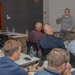 A professional training or business meeting is taking place in a modern conference room. A standing presenter in a gray sweater is addressing a group of approximately 7-8 seated male attendees arranged around white tables. The setting appears to be a corporate workshop or seminar environment.