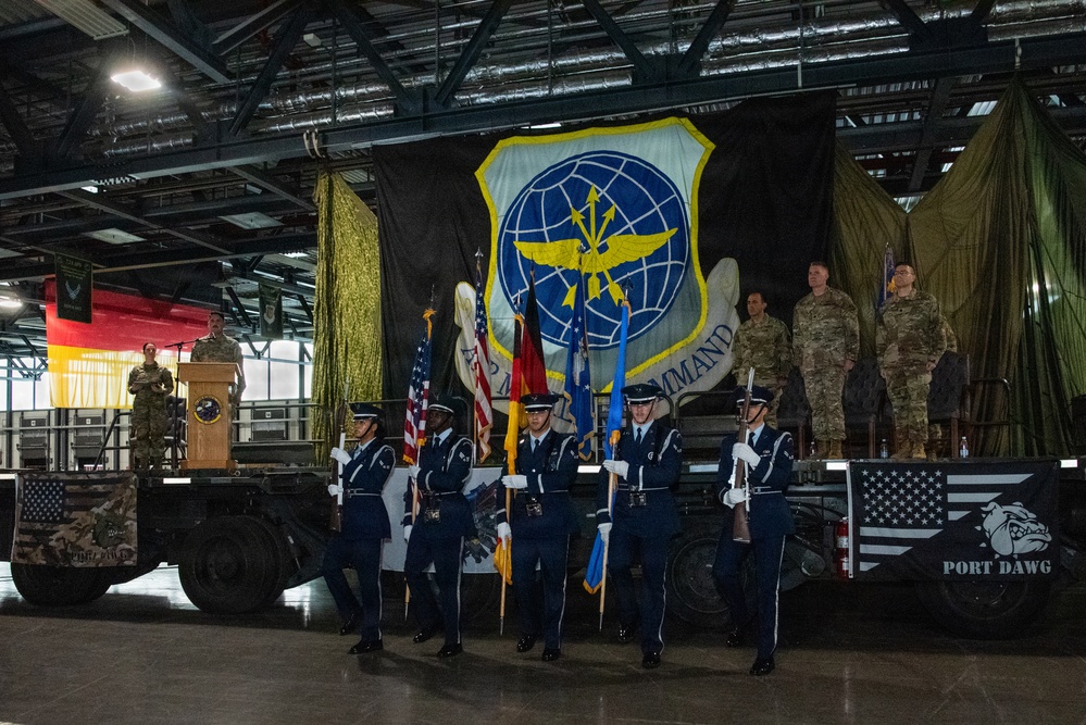 A military ceremony taking place inside a large hangar facility, featuring a Color Guard in U.S. Air Force dress blues presenting flags. A large unit insignia banner is displayed on the backdrop, and military personnel in combat uniforms stand on a raised platform. The event appears to be a formal activation, inactivation, or change of command ceremony.