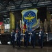 A military ceremony taking place inside a large hangar facility, featuring a Color Guard in U.S. Air Force dress blues presenting flags. A large unit insignia banner is displayed on the backdrop, and military personnel in combat uniforms stand on a raised platform. The event appears to be a formal activation, inactivation, or change of command ceremony.