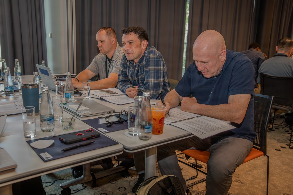 Three men are seated at a conference or workshop table, appearing to be engaged in a meeting or training session. The man in the center is speaking while the others review printed documents. The setting appears to be a modern hotel or conference room.