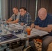 Three men are seated at a conference or workshop table, appearing to be engaged in a meeting or training session. The man in the center is speaking while the others review printed documents. The setting appears to be a modern hotel or conference room.